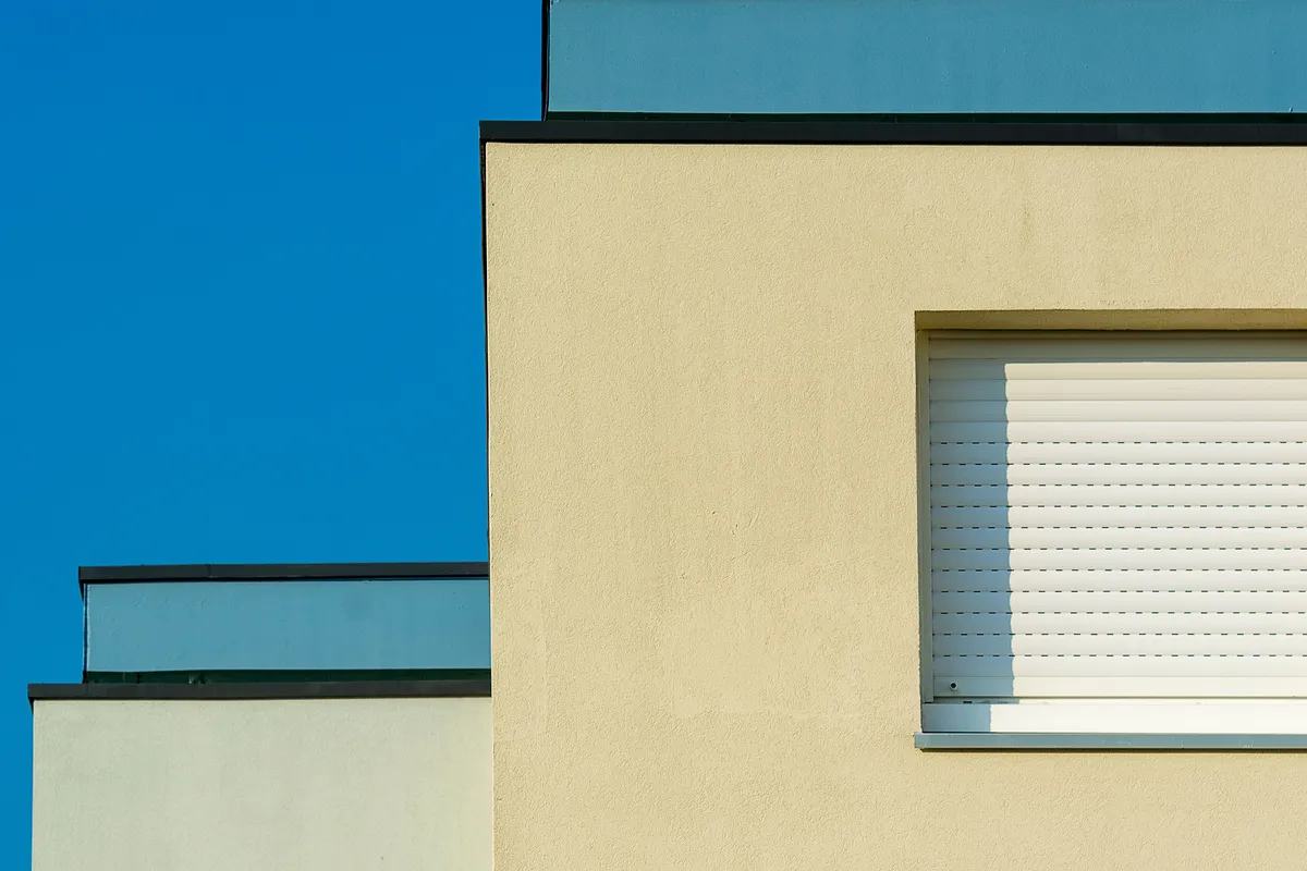 Angle de maison enduit beige à côté d'un mur crème, volet roulant fermé